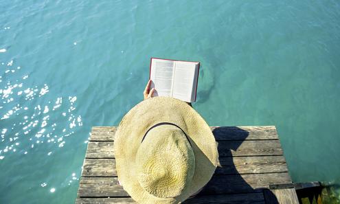 Person mit Strohhut liest auf einem Badesteg ein Buch