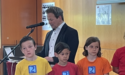 1. Mann spricht bei einem Rednerpult in ein Mikrofon 3 Kinder stehen davor in das Publikum blickend