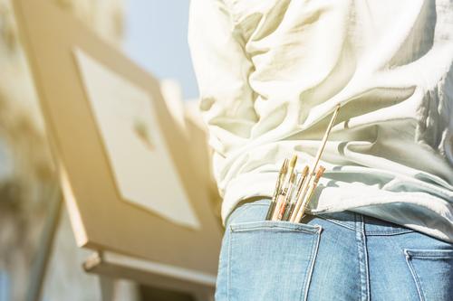 Auf dem Bild steht eine Person vor einer Staffelei und malt im Freien. Sie trägt Jeans und eine helle Bluse. In der Gesäßtasche stecken mehrere Pinsel, und die Szene ist sonnig und hell.