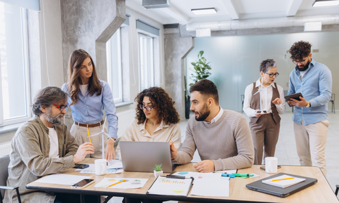 Frauen und Männer stehen und sitzen bei einem Tische und arbeiten zusammen