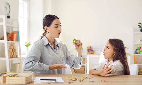 Eine Lehrerin übt Sprechen mit einer Schülerin, beide spitzen die Lippen und gestikulieren dabei