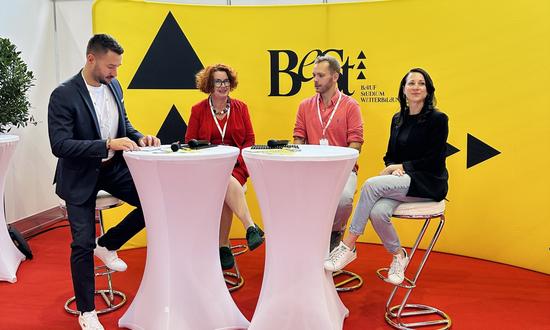 2 Frauen und 1 Mann sitzen auf Barhockern rund um 2 Stehtischen und sind im Gespräch