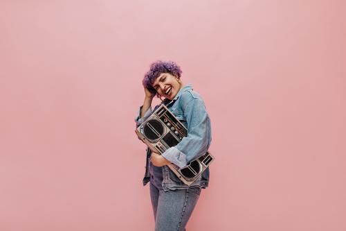 Junge Frau vor rosarotem Hintergrund hält einen Kassettenrekorder, hört Musik und lacht dabei